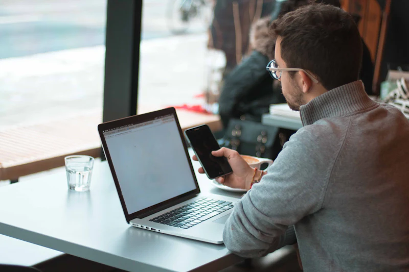 Ondernemer werkt aan een laptop bij het raam en kijkt op zijn telefoon